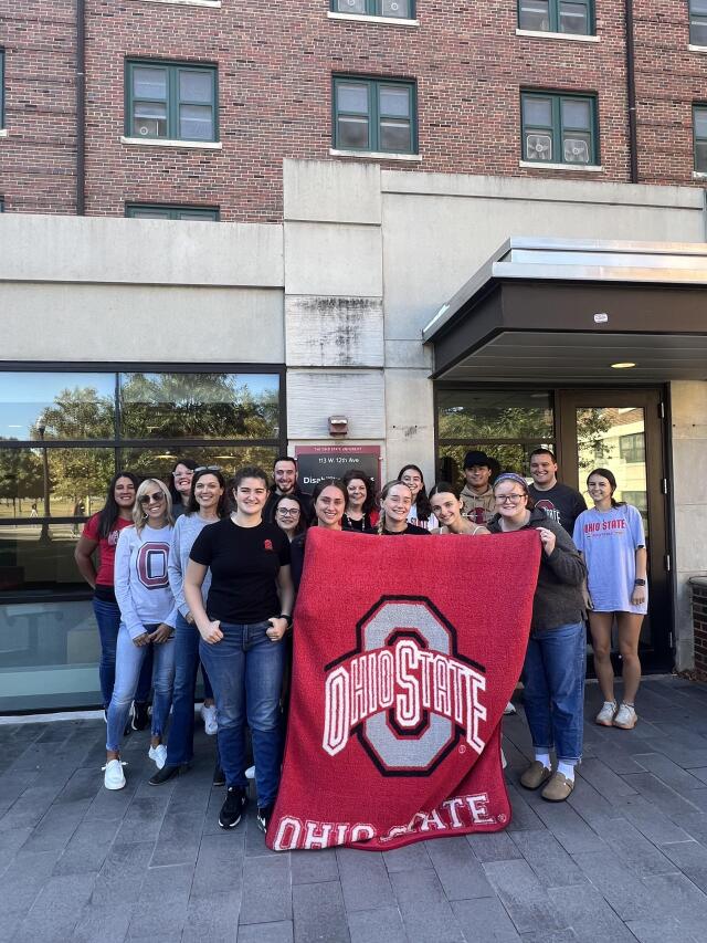 16 Student Life Disability Services professional and student staff members showing off their Buckeye Spirit wearing Scarlet and Gray attire.  They are standing in front of the Baker Hall Disability Services sign and they are holding up a scarlet blanket de