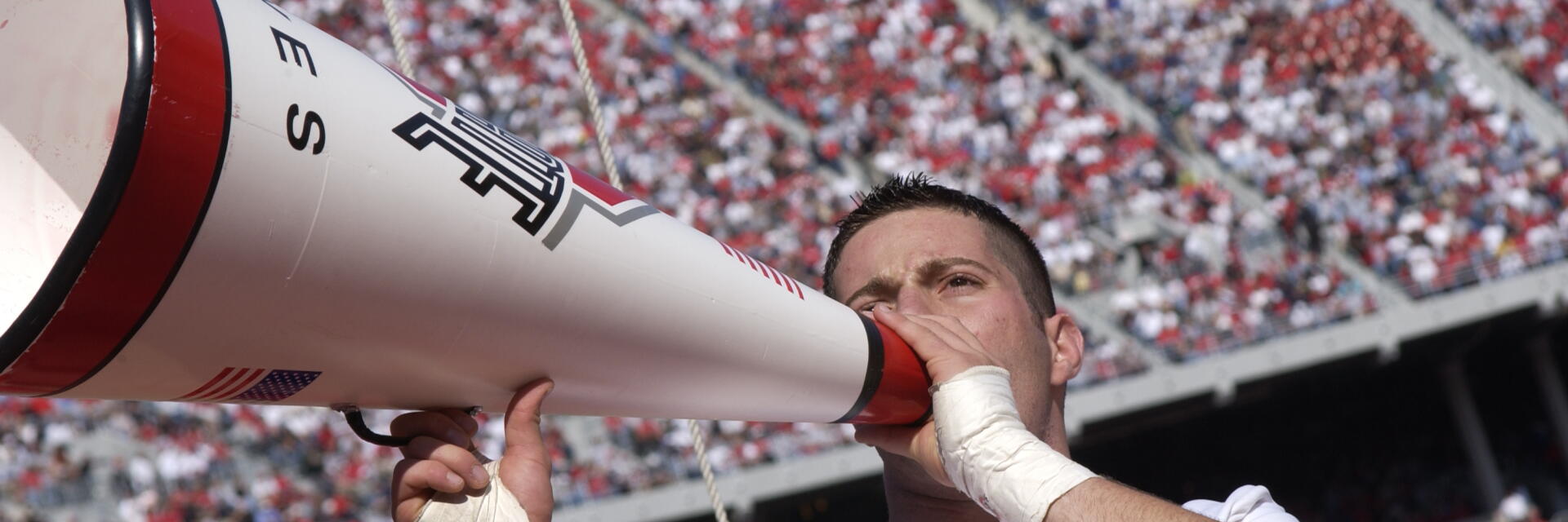 An Ohio State male cheerleader in uniform, shouting through a megaphone to a Stadium crowd.