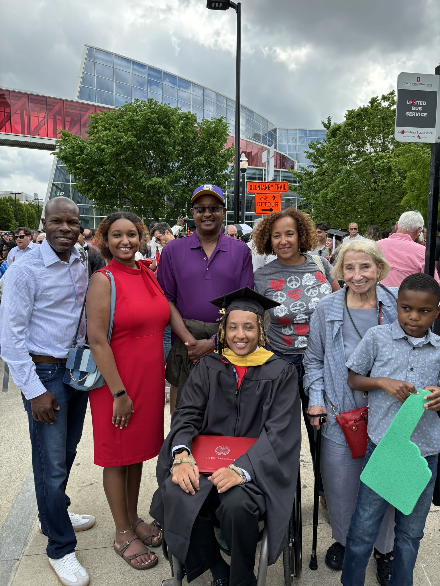 Justin Lennon, a black man in a wheelchair, wearing graduation robes, surrounded by 6 supporters.