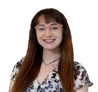 Carly Taylor, a smiling white woman with long brown hair.  She is wearing a black and white floral blouse and light brown glasses.