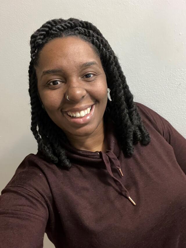 Jamonae Scarborough - A smiling Black woman with shoulder length twists, wearing brown top and a silver hoop nose ring.