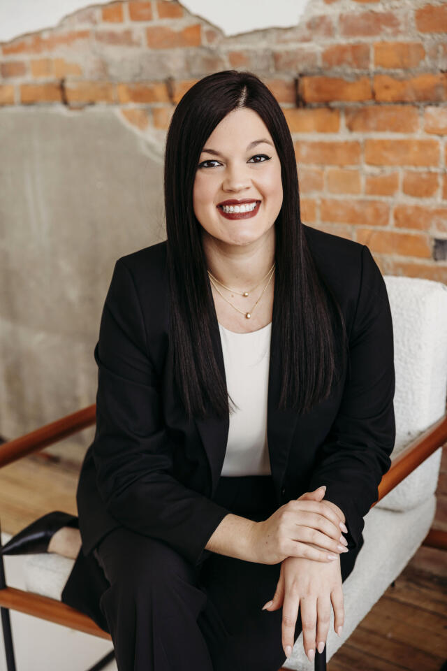 A white woman with long, straight, brown hair, wearing a black blazer over a white blouse.