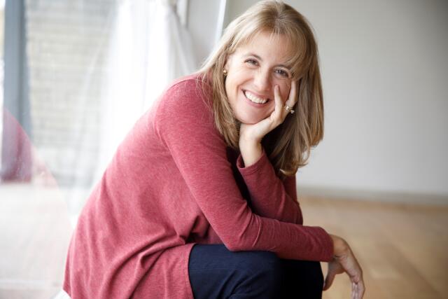 A smiling white woman wearing a mauve sweater and navy pants, with shoulder length blond hair.