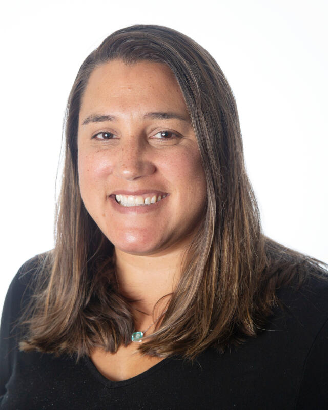 A woman with long brown hair wearing a black, v-neck blouse and a necklace.