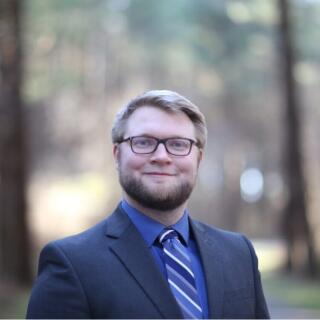 "A smiling, blonde, White man wearing a suit, collared shirt and striped tie."