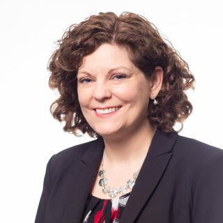 A white woman with chin length curly brown hair, wearing a blazer and blouse.