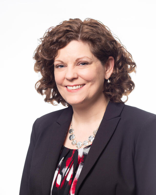 A white woman with chin length curly brown hair, wearing a blazer and blouse.