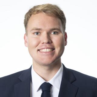 A smiling white man with blonde hair, wearing a white shirt, blue suit coat, and tie.