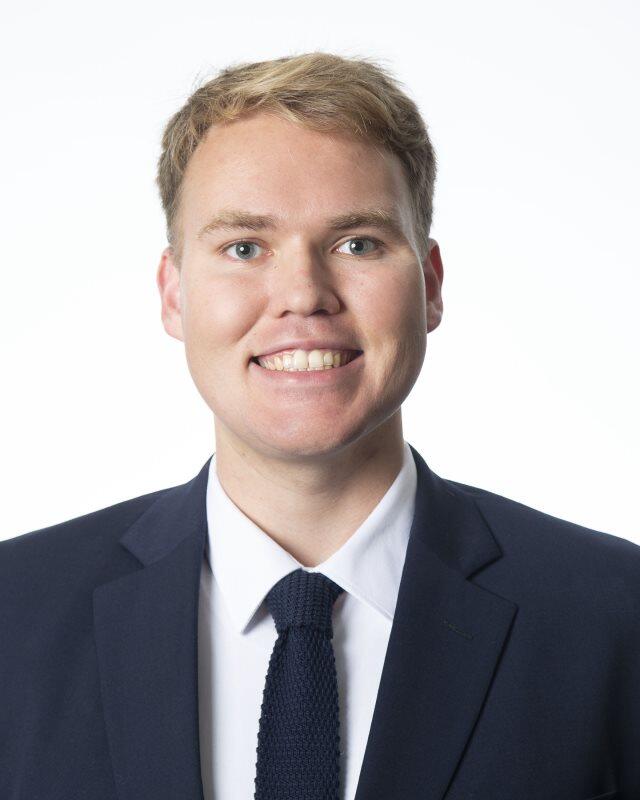 A smiling white man with blonde hair, wearing a white shirt, blue suit coat, and tie.
