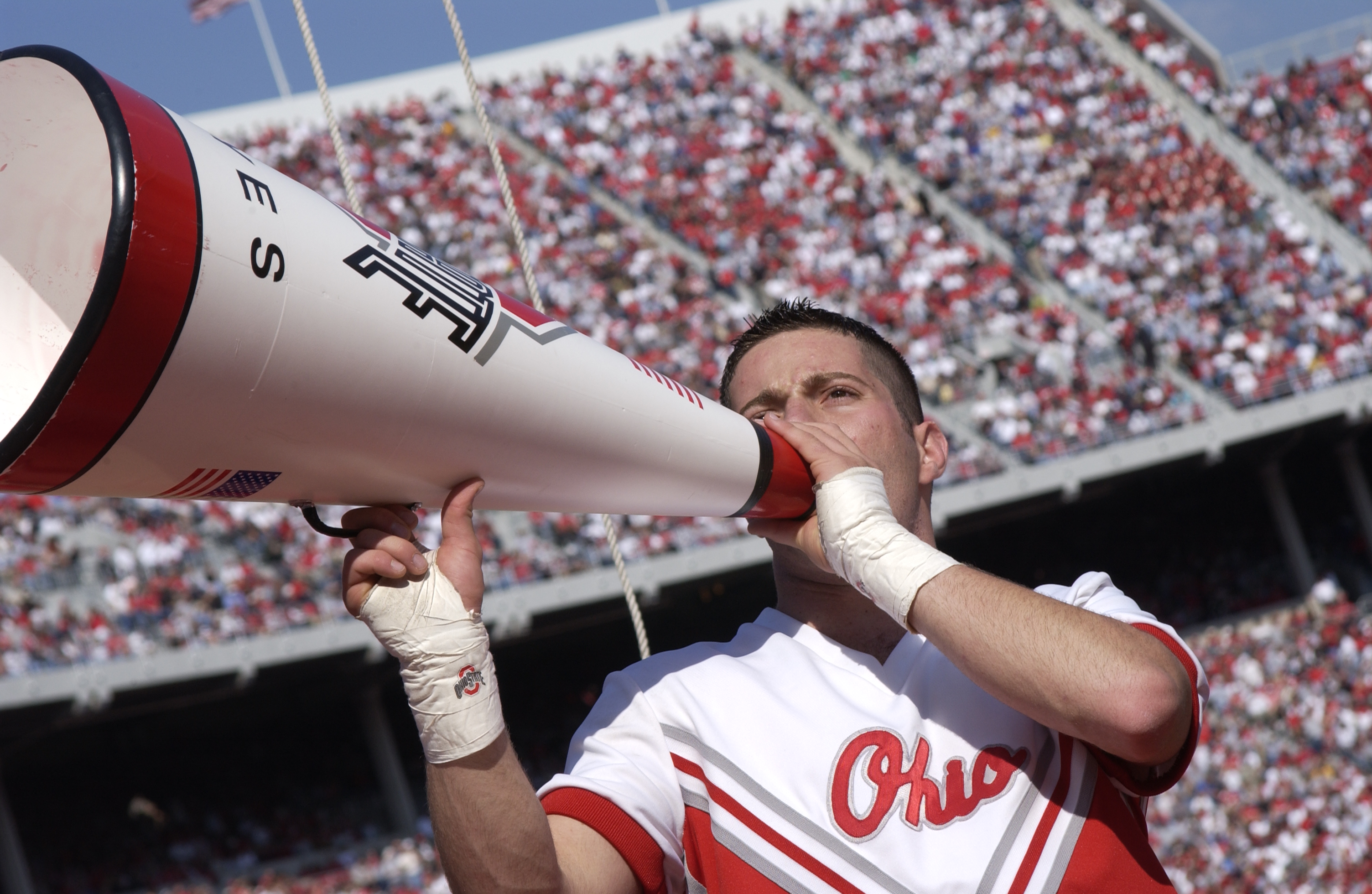 An Ohio State male cheerleader in uniform, shouting through a megaphone to a Stadium crowd.