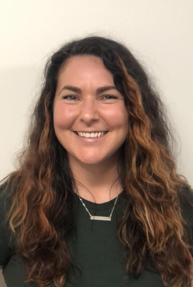 A smiling white woman with long, wavy brown hair wearing a green t-shirt and silver necklace.