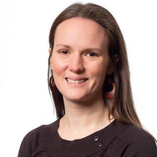 A white woman with straight brown hair wearing earrings and a black scoop neck blouse.