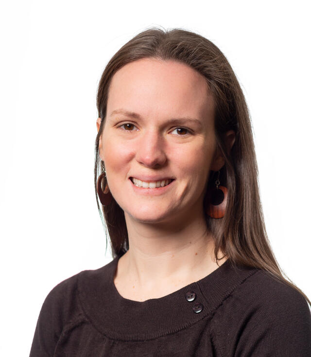 A white woman with straight brown hair wearing earrings and a black scoop neck blouse.
