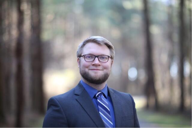 A smiling, blonde, White man wearing a suit, collared shirt and striped tie."