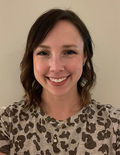 A smiling white woman with shoulder length brown hair, wearing an animal print blouse.