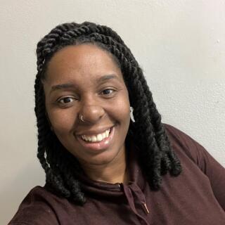A smiling black woman with shoulder length twists, wearing brown top and a nose ring.