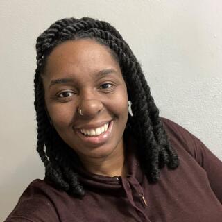 Jamonae Scarborough - A Black woman with shoulder length twists, wearing brown top and a nose ring.