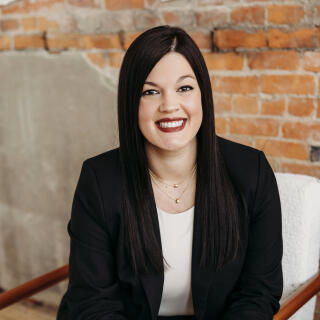 A white woman with long, straight, brown hair, wearing a black blazer over a white blouse.