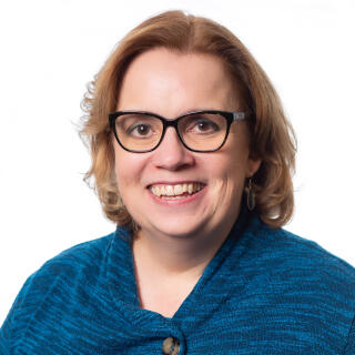 A smiling white woman with dark framed glasses and wavy light brown hair wearing a blue sweater.