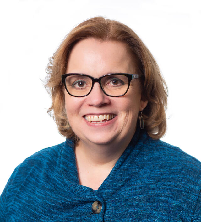 A smiling white woman with dark framed glasses and wavy light brown hair wearing a blue sweater.