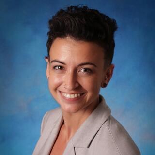 A smiling woman with short, dark, wavy hair, wearing a gray suit jacket with a black blouse.