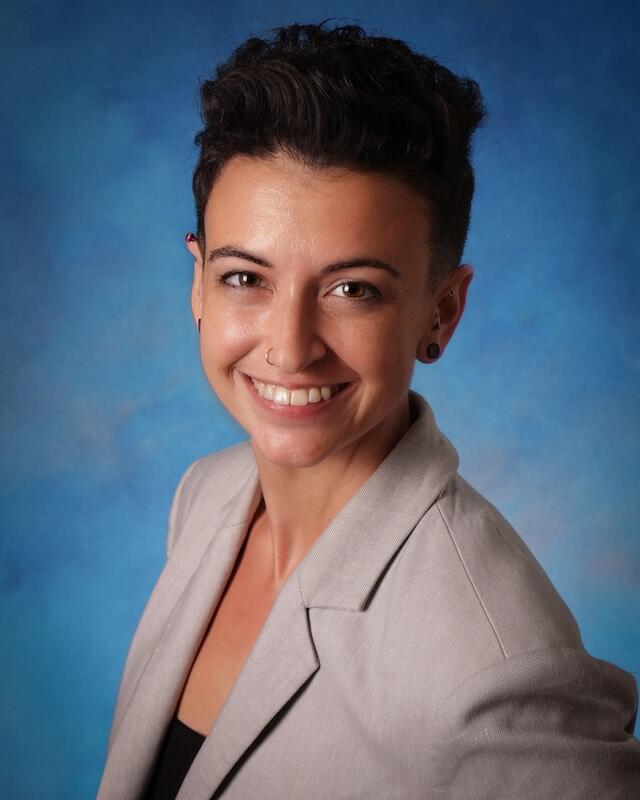 A smiling woman with short, dark, wavy hair, wearing a gray suit jacket with a black blouse.