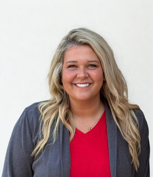 A smiling white woman with long blonde hair, wearing a red dress with a gray blazer
