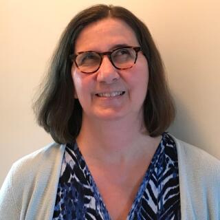 A smiling white woman with brown hair wearing a blouse, a sweater, and glasses.