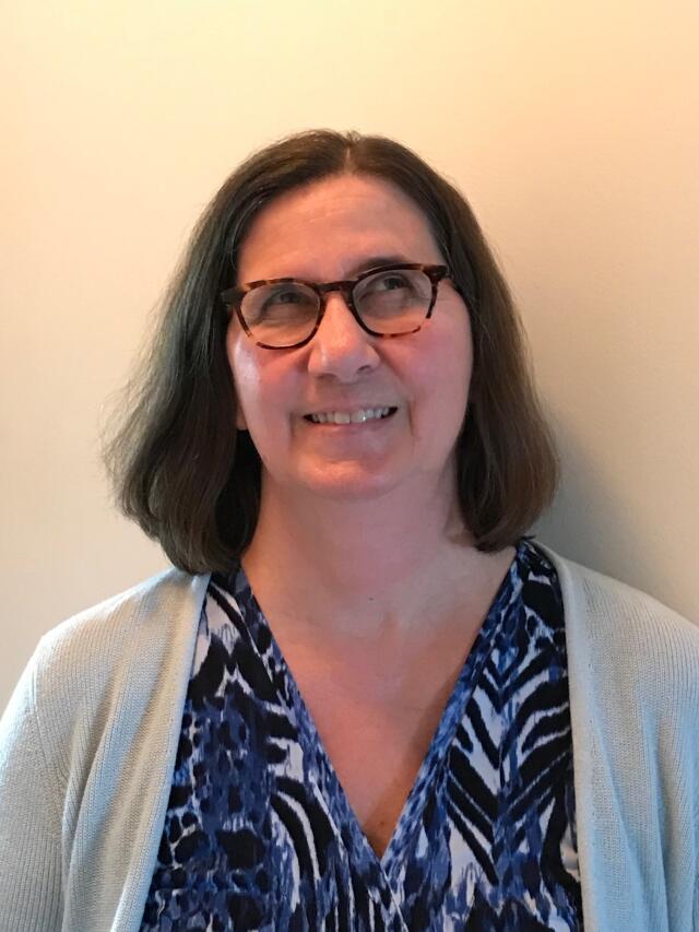 A smiling white woman with brown hair wearing a blouse, a sweater, and glasses.