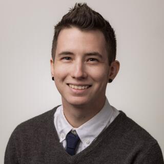 Robbie Long, smiling in a shirt and tie with a sweater. They have short hair and black earrings.