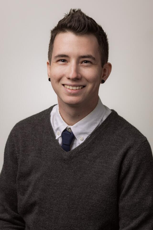 Smiling in a shirt and tie with a sweater. They have short hair and black earrings.