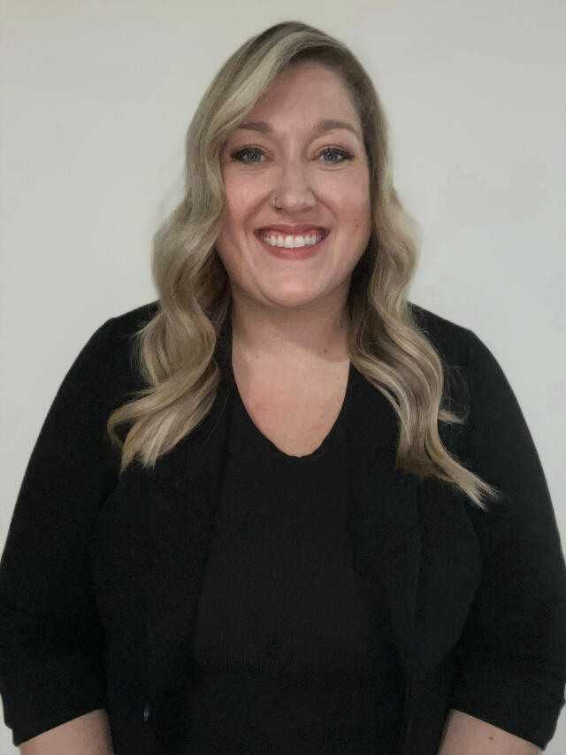 A smiling white woman with wavy blonde hair, wearing a black top and blazer.