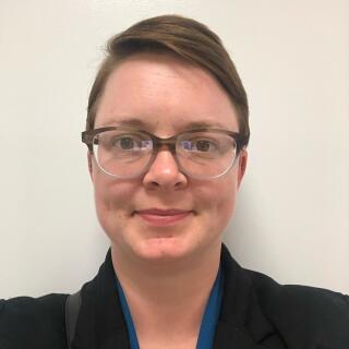 a smiling white woman with short brown hair and glasses, wearing a blue blouse and black blazer.