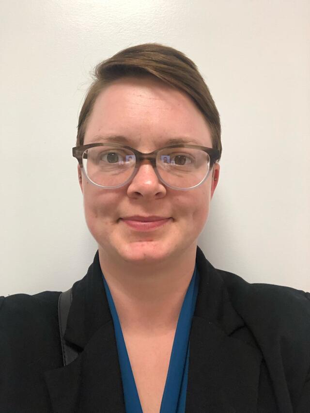 a smiling white woman with short brown hair and glasses, wearing a blue blouse and black blazer.