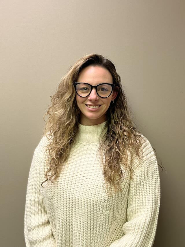 A white woman with long curly light brown hair, wearing glasses and a turtleneck.