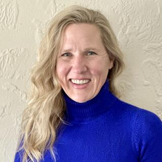 A smiling white woman with wavy blond hair, parted on the side, wearing a blue turtleneck.