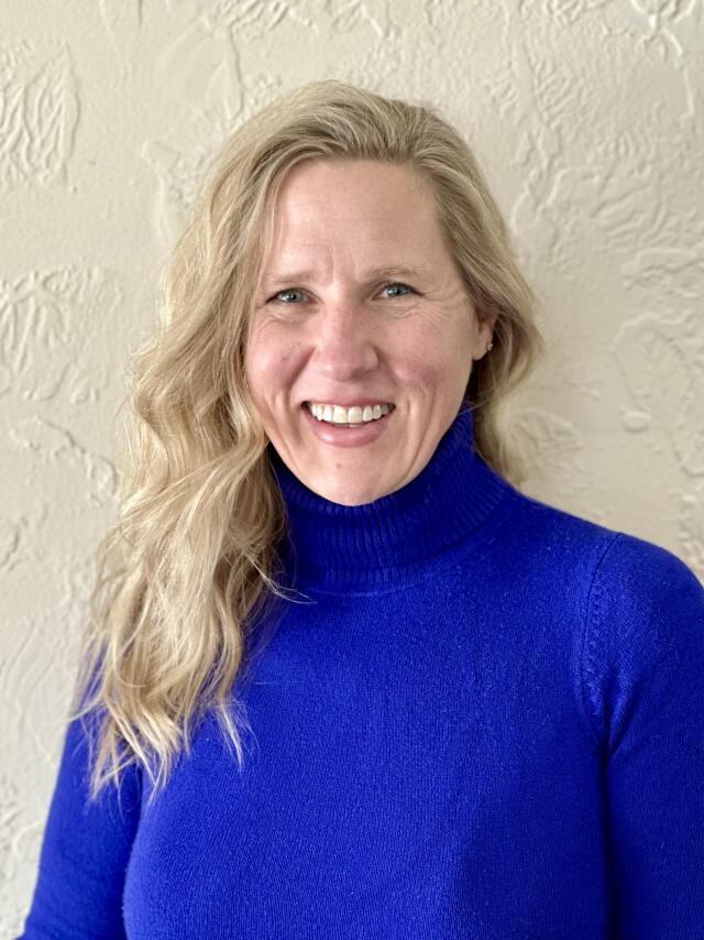 A smiling white woman with wavy blond hair, parted on the side, wearing a blue turtleneck.